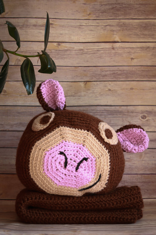 A crocheted cow head pillow sits on top of a matching blanket in front of a weathered wood background with a plant visible in the upper left corner of the frame.