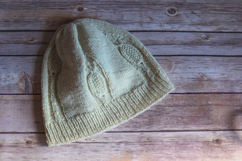 An off-white knitted hat with textural patterns resembling teasel plants lays flat against a weathered wood background.